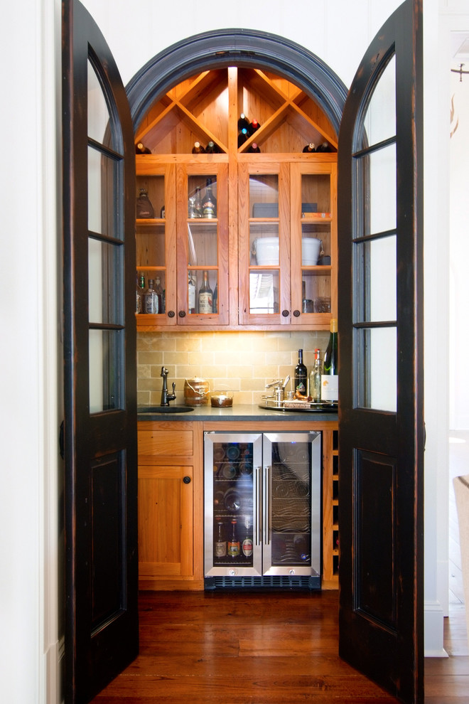Example of a small classic single-wall dark wood floor wet bar design in New Orleans with shaker cabinets, medium tone wood cabinets and beige backsplash