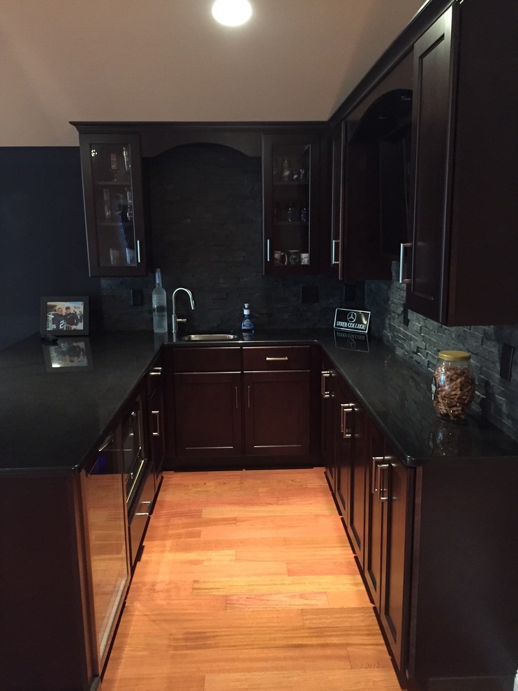 Inspiration for a medium sized traditional u-shaped wet bar in Philadelphia with medium hardwood flooring, a submerged sink, recessed-panel cabinets, dark wood cabinets, composite countertops, grey splashback, stone tiled splashback and brown floors.
