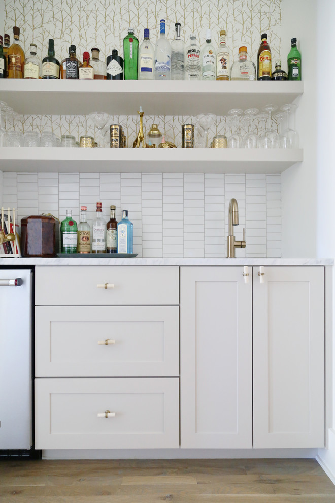 Bild på en mellanstor funkis linjär hemmabar med vask, med en undermonterad diskho, skåp i shakerstil, vita skåp, marmorbänkskiva, vitt stänkskydd, stänkskydd i porslinskakel och ljust trägolv