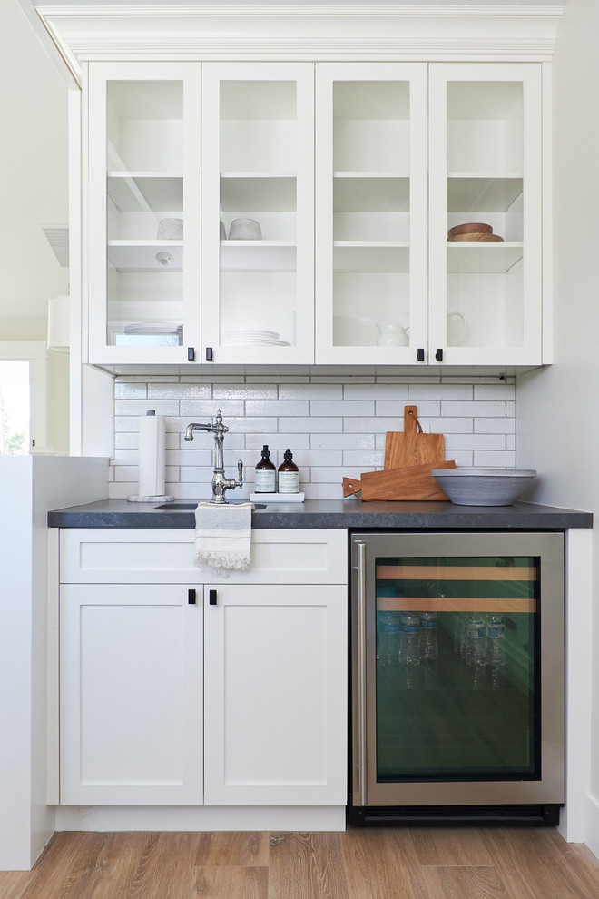 Example of a small beach style single-wall ceramic tile and brown floor wet bar design in San Diego with an undermount sink, shaker cabinets, white cabinets, quartz countertops, white backsplash, ceramic backsplash and black countertops