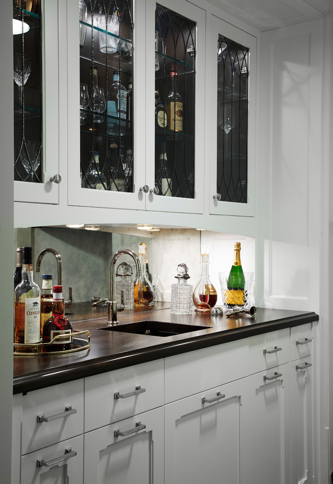 Klassisk inredning av en mellanstor svarta linjär svart hemmabar med vask, med en undermonterad diskho, luckor med glaspanel, vita skåp, bänkskiva i koppar, spegel som stänkskydd, mörkt trägolv och brunt golv