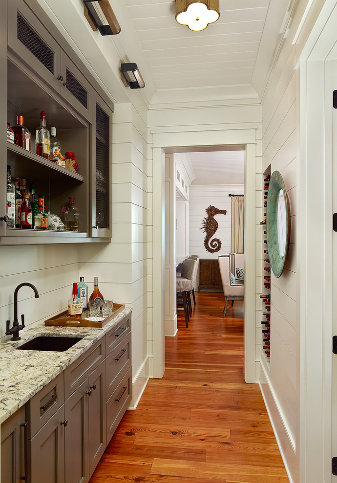 Inspiration for a tropical single-wall medium tone wood floor and brown floor wet bar remodel in Charleston with an undermount sink, recessed-panel cabinets, gray cabinets and white backsplash