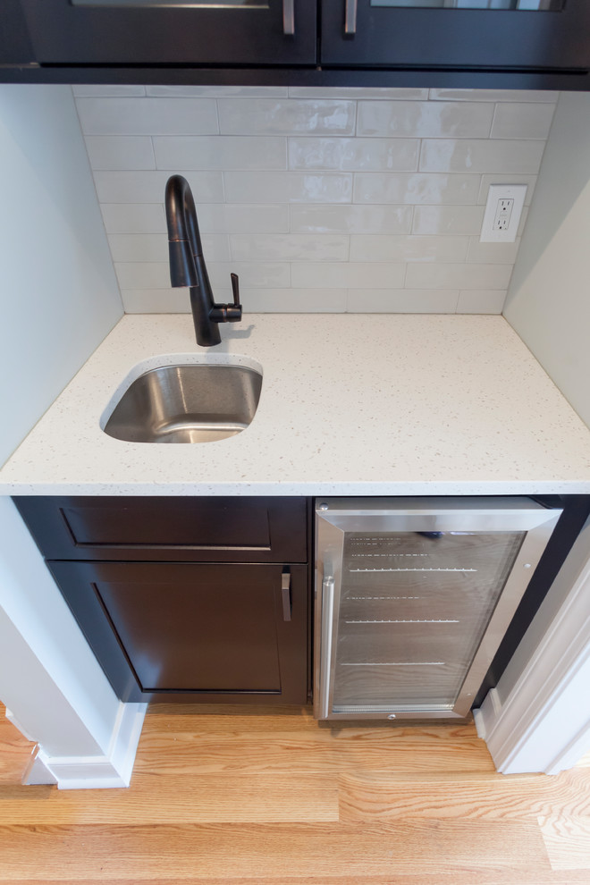 Photo of a small classic single-wall wet bar in Philadelphia with a submerged sink, shaker cabinets, dark wood cabinets, engineered stone countertops, white splashback, metro tiled splashback, light hardwood flooring, brown floors and white worktops.