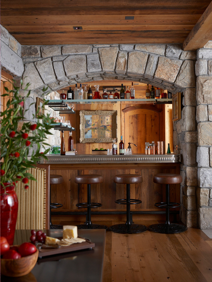 Cette image montre un bar de salon rustique avec des tabourets, un placard sans porte et un sol en bois brun.