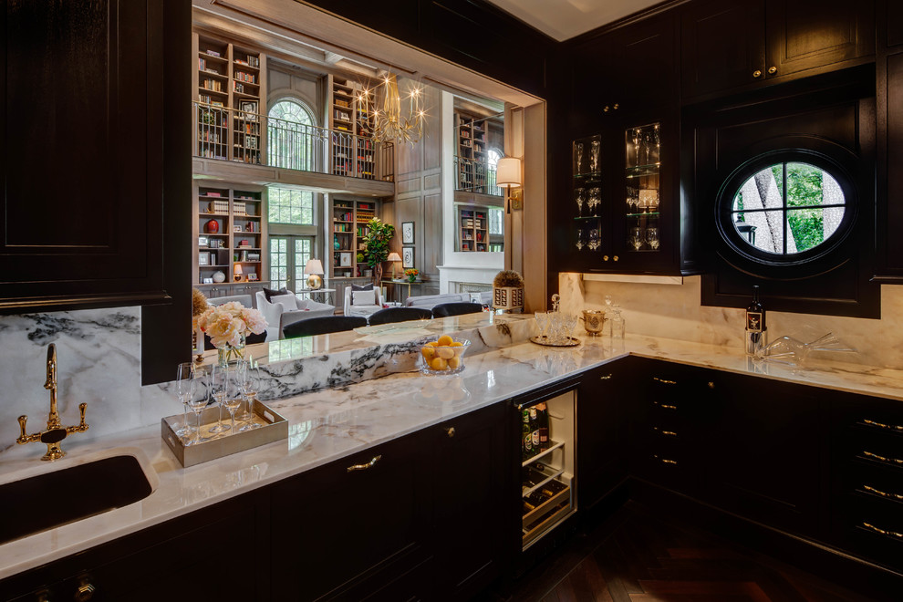 Diseño de bar en casa con barra de bar clásico renovado grande con fregadero bajoencimera, armarios con paneles empotrados, puertas de armario negras, encimera de mármol, salpicadero blanco, salpicadero de mármol, suelo de madera oscura y encimeras blancas