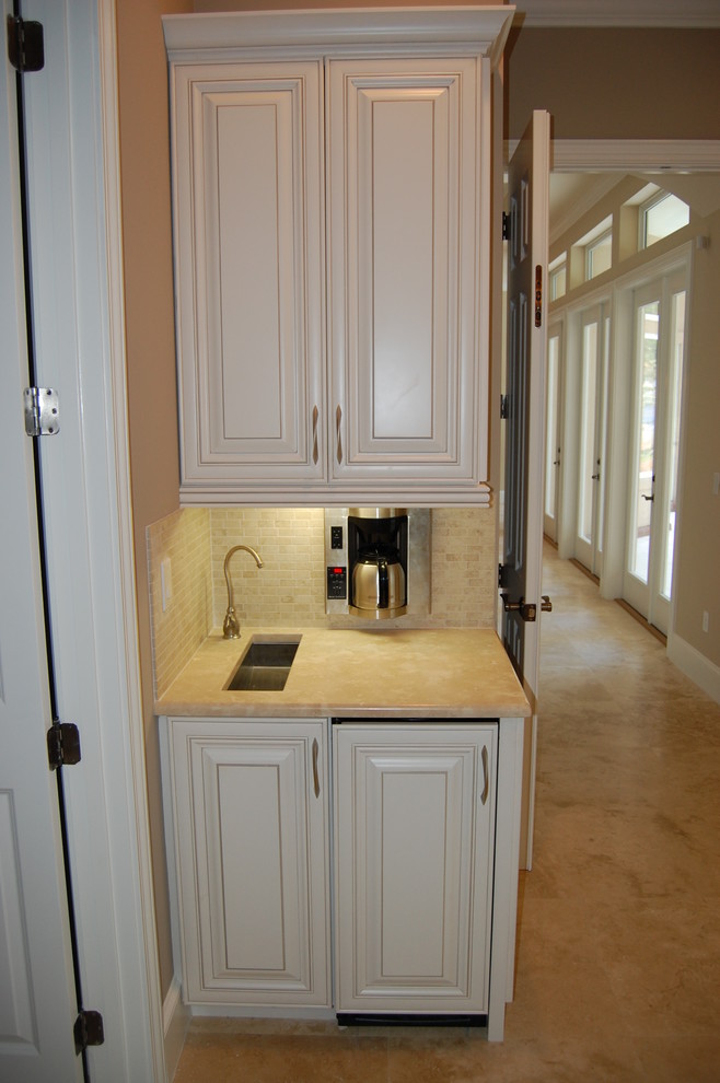 Inspiration for a small timeless single-wall travertine floor wet bar remodel in Jacksonville with an undermount sink, raised-panel cabinets, white cabinets, limestone countertops, beige backsplash and stone tile backsplash