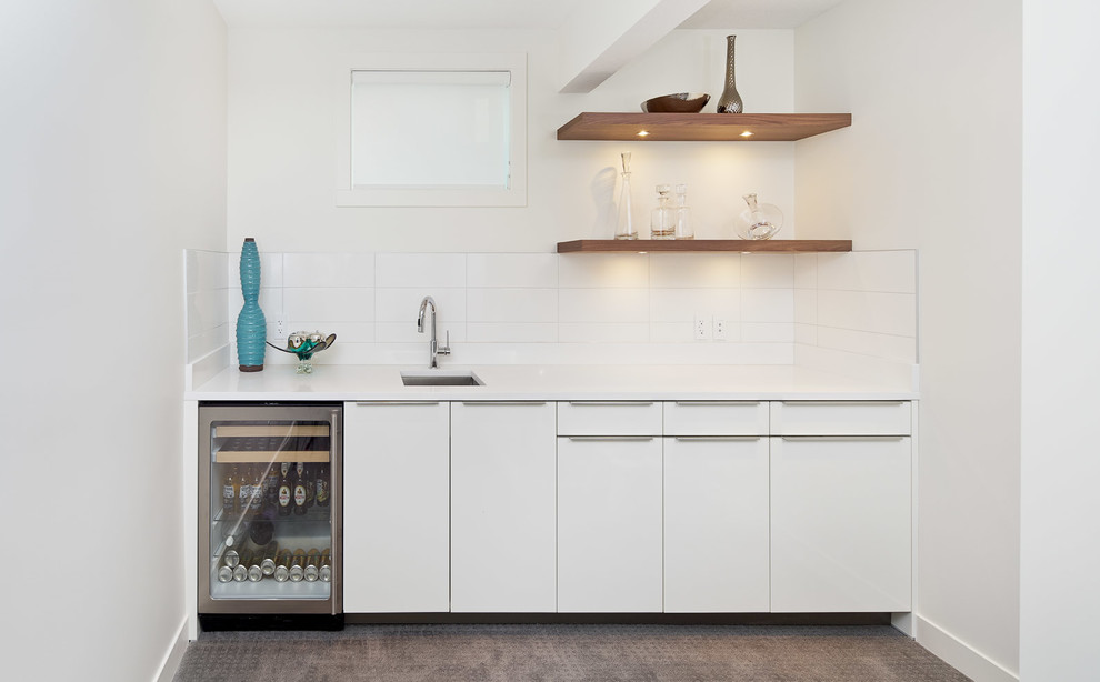 This is an example of a small contemporary single-wall wet bar in Calgary with a submerged sink, flat-panel cabinets, white cabinets, engineered stone countertops, white splashback, carpet and metro tiled splashback.