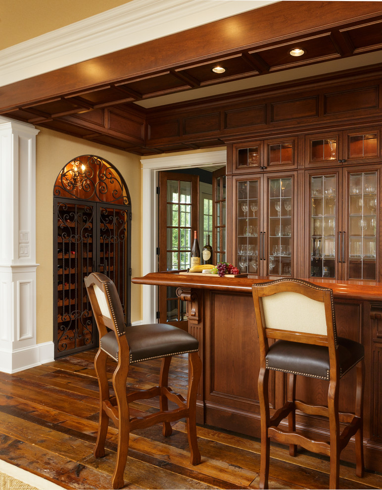 Modelo de bar en casa con barra de bar tradicional con armarios tipo vitrina, puertas de armario de madera oscura y suelo de madera en tonos medios