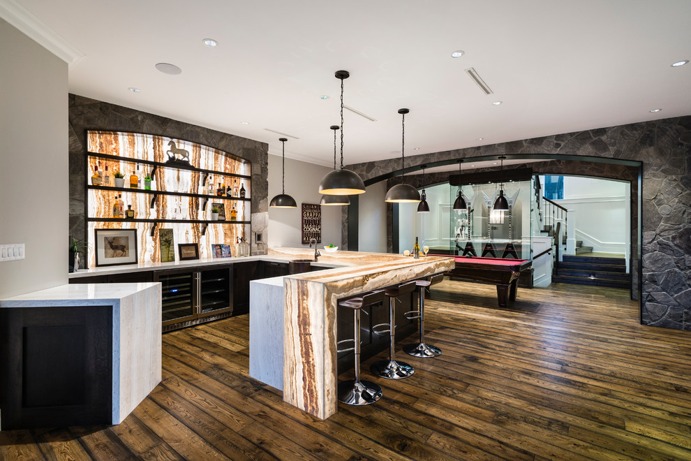 Example of a huge mountain style u-shaped medium tone wood floor and brown floor wet bar design in Vancouver with a drop-in sink, shaker cabinets, dark wood cabinets, onyx countertops, beige backsplash, stone slab backsplash and beige countertops