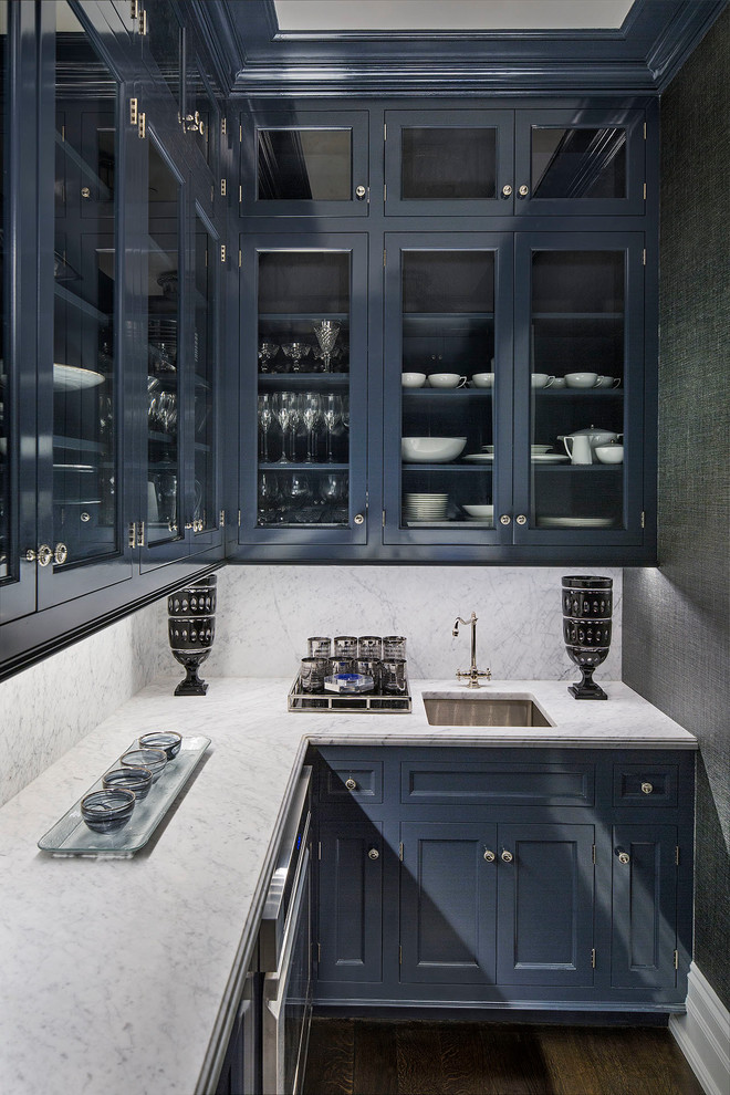 Inredning av en klassisk mellanstor l-formad hemmabar med vask, med en undermonterad diskho, luckor med glaspanel, blå skåp, marmorbänkskiva, vitt stänkskydd, stänkskydd i marmor, mörkt trägolv och brunt golv