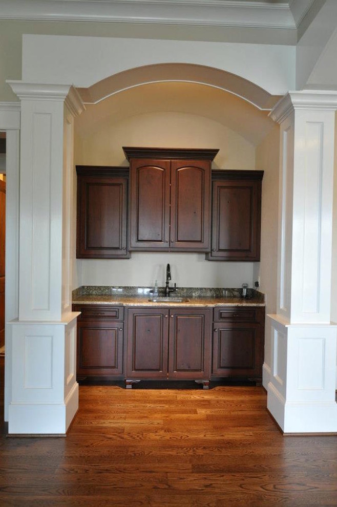 This is an example of a small traditional single-wall wet bar in Other with raised-panel cabinets, dark wood cabinets, granite worktops, medium hardwood flooring, brown floors and a submerged sink.