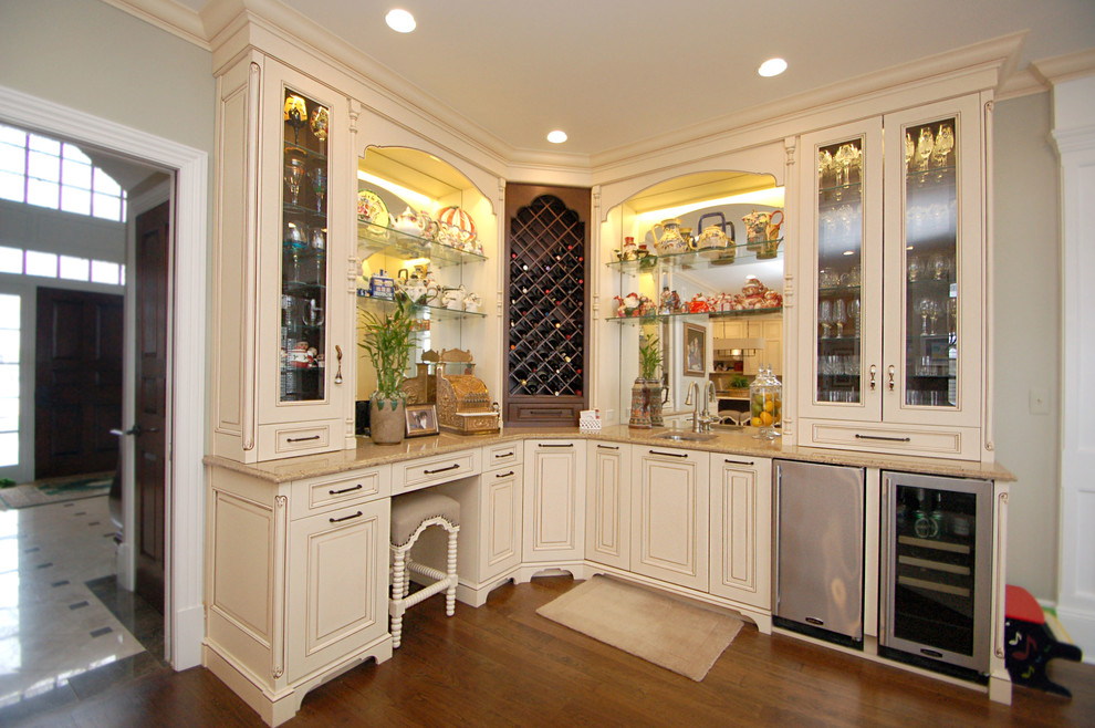 This is an example of a large traditional l-shaped wet bar in Newark with a submerged sink, raised-panel cabinets, white cabinets, engineered stone countertops, white splashback, mirror splashback and medium hardwood flooring.