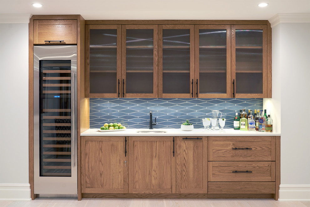 Photo of a nautical single-wall wet bar in New York with a submerged sink, glass-front cabinets, medium wood cabinets, blue splashback, light hardwood flooring, beige floors and white worktops.