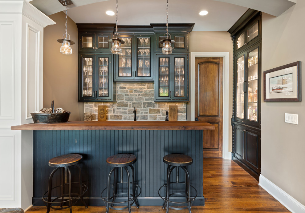 Imagen de bar en casa con barra de bar clásico de tamaño medio con armarios tipo vitrina, puertas de armario negras, encimera de madera, salpicadero de azulejos de piedra y suelo de madera en tonos medios