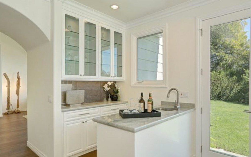 Photo of a small modern galley wet bar in Los Angeles with a submerged sink, raised-panel cabinets, white cabinets, marble worktops, beige splashback, porcelain splashback and light hardwood flooring.