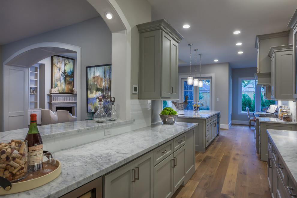 Photo of an expansive traditional galley wet bar in Houston with no sink, recessed-panel cabinets, grey cabinets, marble worktops, white splashback, metro tiled splashback, light hardwood flooring, brown floors and grey worktops.