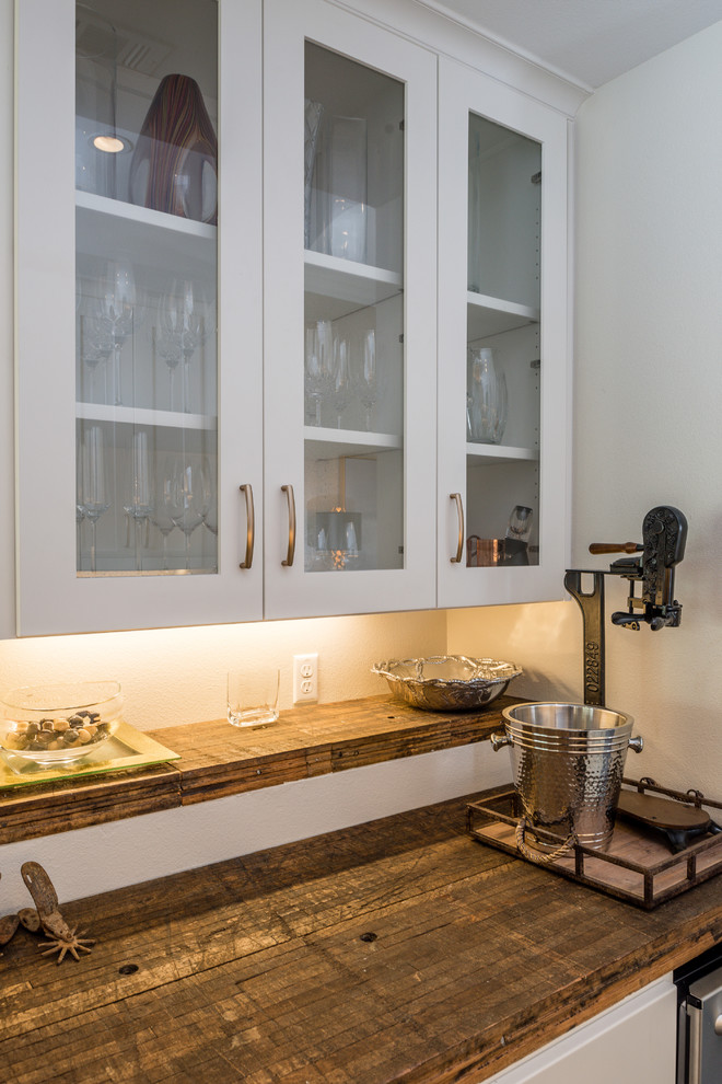 This is an example of a small traditional galley home bar in Austin with glass-front cabinets, white cabinets, wood worktops and white splashback.