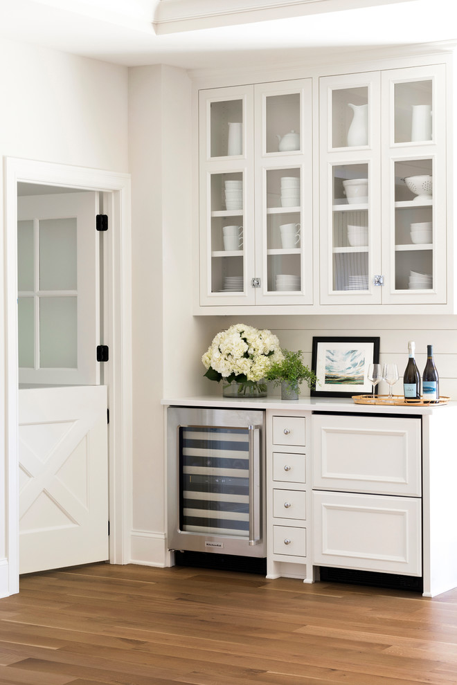 Diseño de bar en casa lineal costero sin pila con armarios tipo vitrina, puertas de armario blancas, encimeras blancas y suelo de madera clara