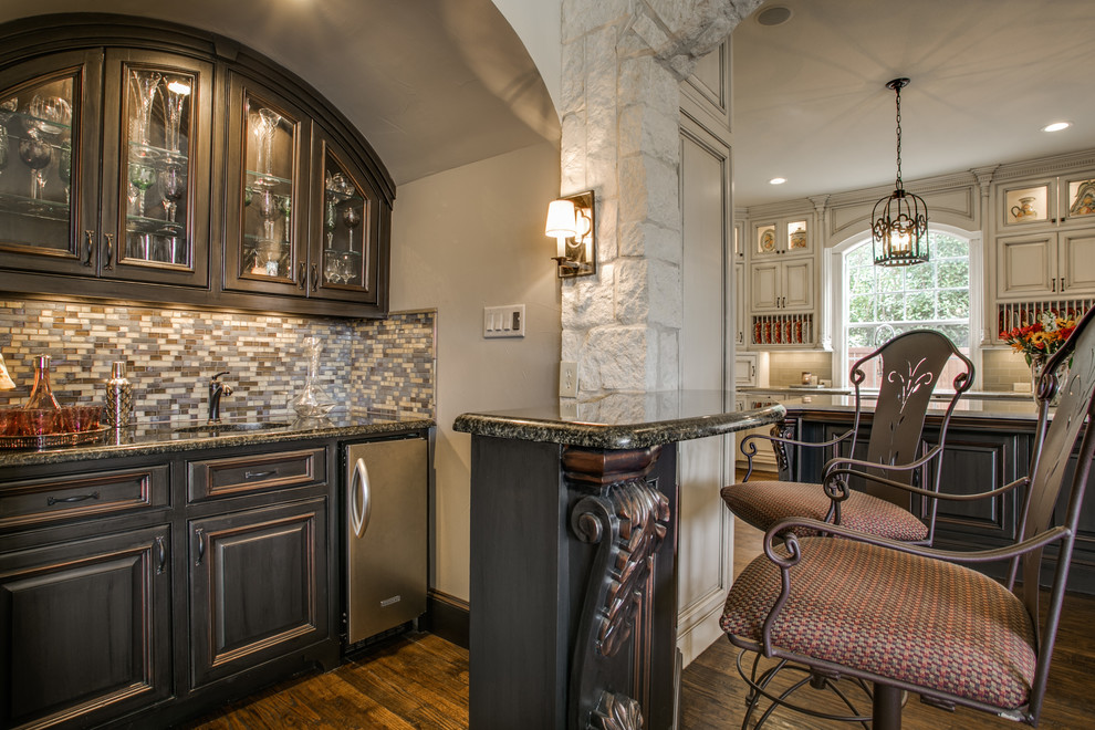 Inspiration for a mid-sized timeless galley dark wood floor and brown floor seated home bar remodel in Dallas with an undermount sink, raised-panel cabinets, dark wood cabinets, granite countertops, multicolored backsplash and matchstick tile backsplash
