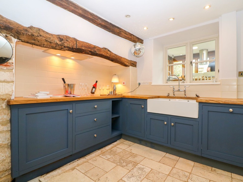 Photo of a small farmhouse l-shaped home bar in Gloucestershire with a built-in sink, shaker cabinets, blue cabinets, wood worktops, white splashback, metro tiled splashback, limestone flooring and beige floors.