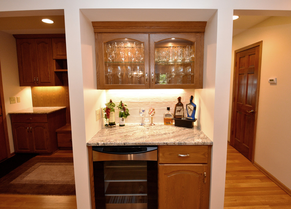 Small traditional single-wall wet bar in Minneapolis with no sink, glass-front cabinets, light wood cabinets, granite worktops, white splashback, porcelain splashback, light hardwood flooring, brown floors and multicoloured worktops.