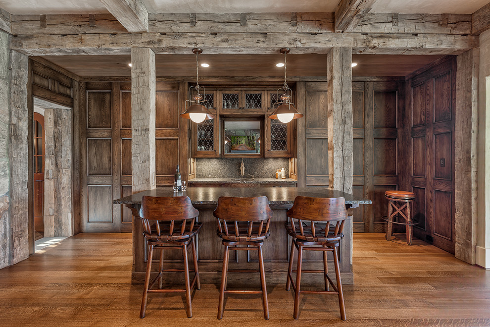 Inspiration pour un grand bar de salon parallèle chalet en bois foncé avec un plan de travail en granite, un sol en bois brun, une crédence grise, des tabourets, un évier encastré, un placard à porte vitrée, une crédence en dalle de pierre et un sol marron.