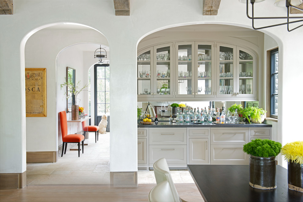 This is an example of a traditional single-wall wet bar in New York with glass-front cabinets, beige cabinets, mirror splashback, light hardwood flooring and beige floors.