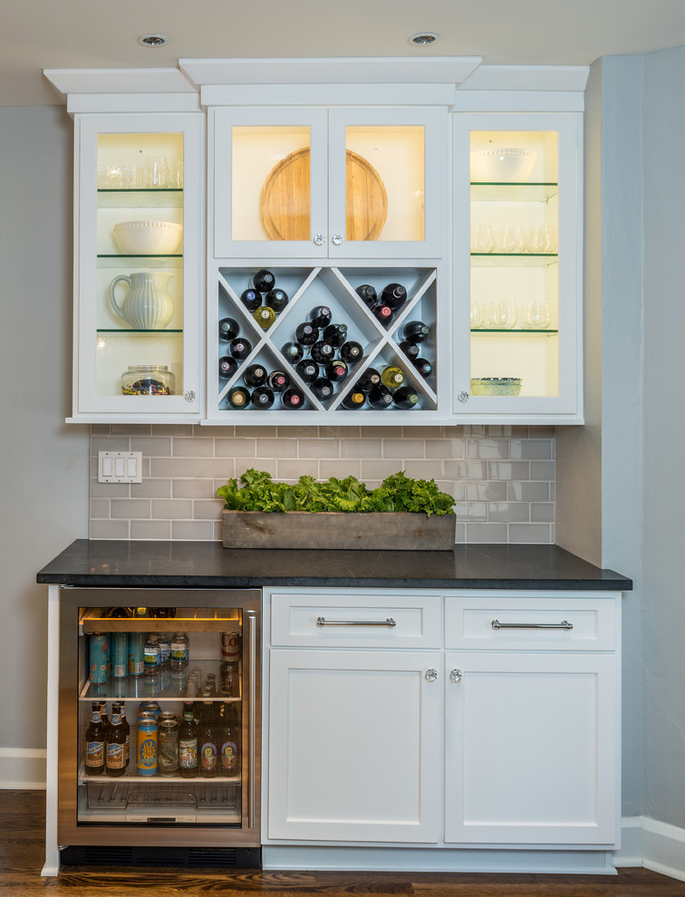 Home bar - transitional single-wall medium tone wood floor home bar idea in Indianapolis with no sink, glass-front cabinets, white cabinets, gray backsplash, subway tile backsplash and black countertops