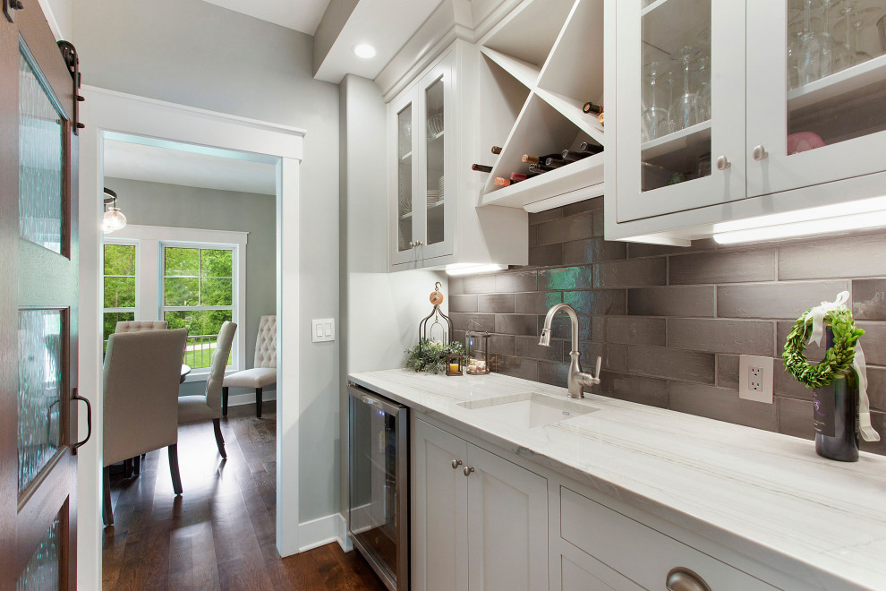 Example of a mid-sized cottage single-wall dark wood floor and brown floor wet bar design in Kansas City with an undermount sink, shaker cabinets, white cabinets, white countertops, quartzite countertops, gray backsplash and glass tile backsplash