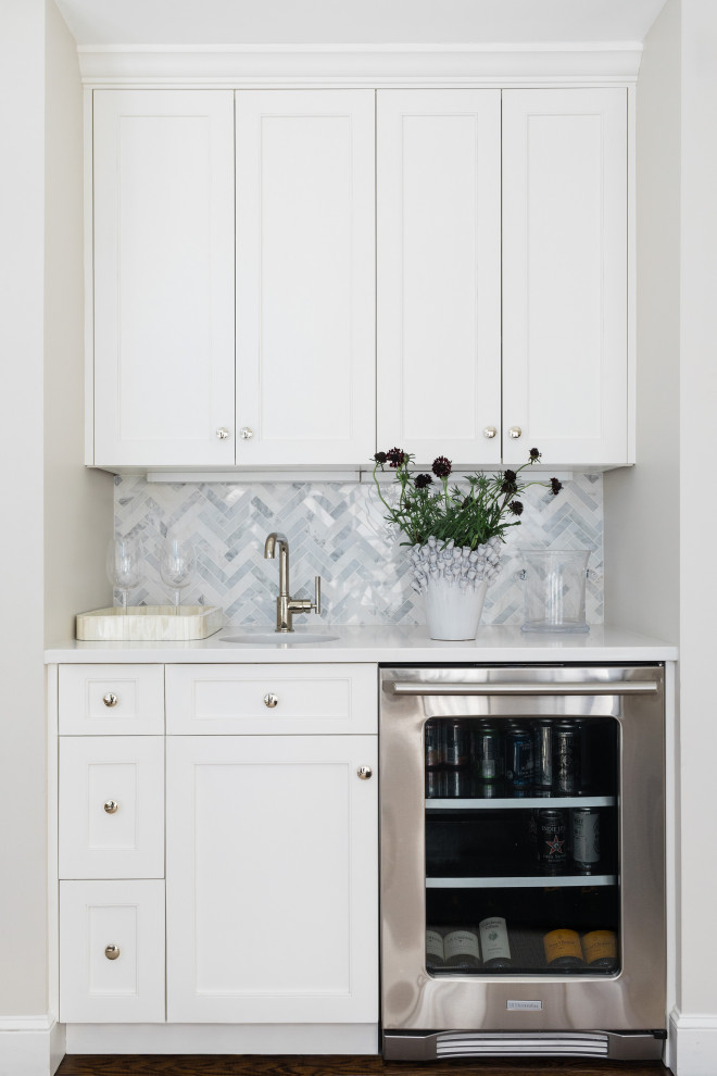 This is an example of a small classic single-wall wet bar in Boston with a submerged sink, recessed-panel cabinets, white cabinets, engineered stone countertops, white splashback, marble splashback, medium hardwood flooring, brown floors and white worktops.