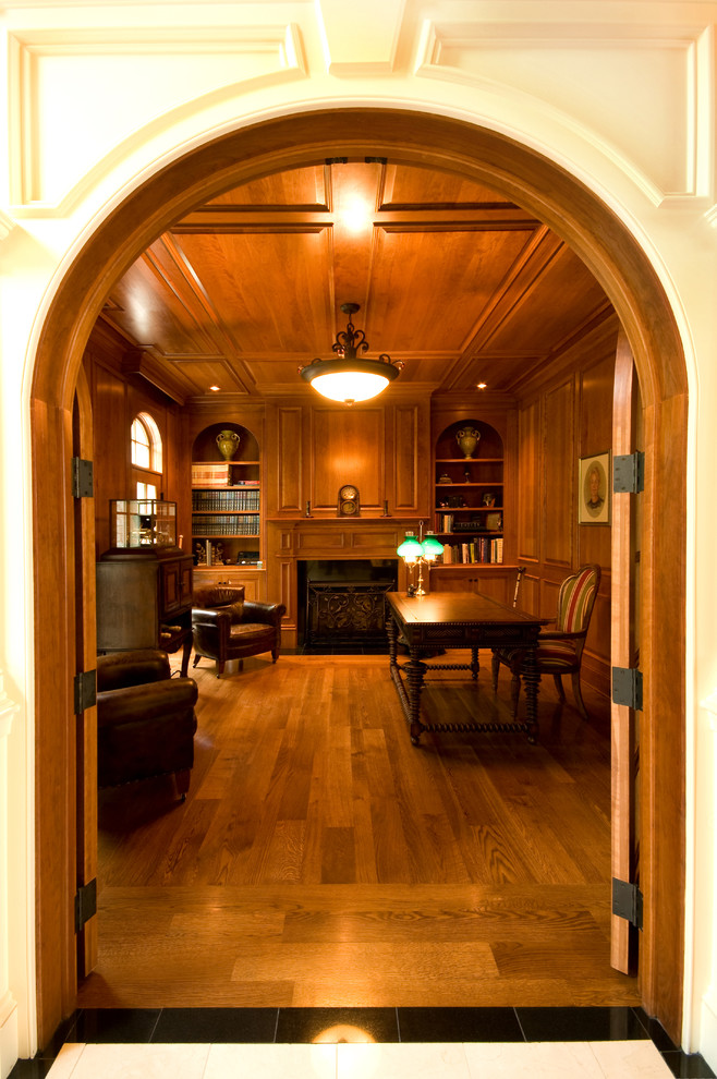 Bild på ett stort vintage arbetsrum, med ett bibliotek, bruna väggar, mörkt trägolv, en standard öppen spis, en spiselkrans i trä, ett fristående skrivbord och brunt golv