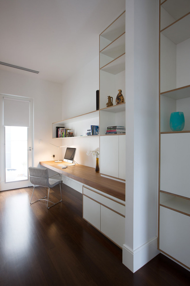 Diseño de despacho contemporáneo con paredes blancas, suelo de madera oscura, escritorio empotrado y suelo marrón