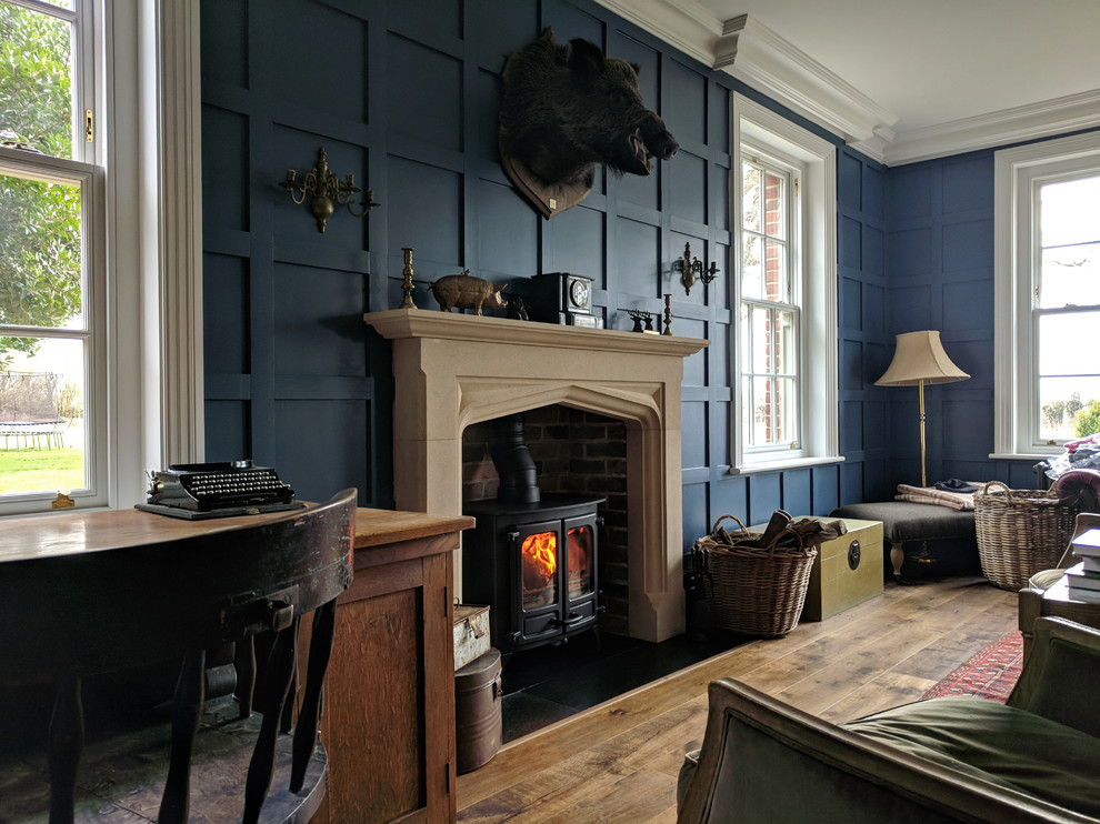 Exempel på ett mellanstort klassiskt hemmabibliotek, med blå väggar, mellanmörkt trägolv, en öppen vedspis, en spiselkrans i sten, ett fristående skrivbord och brunt golv