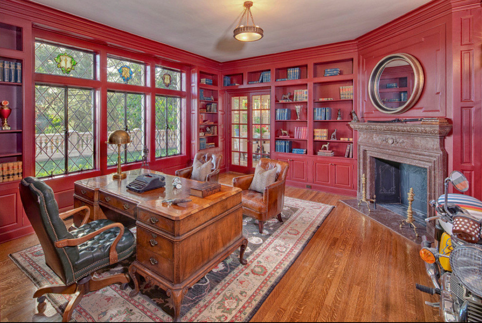 Cette photo montre un grand bureau chic avec un mur rouge, un sol en bois brun, une cheminée d'angle, un manteau de cheminée en pierre, un bureau indépendant et un sol marron.