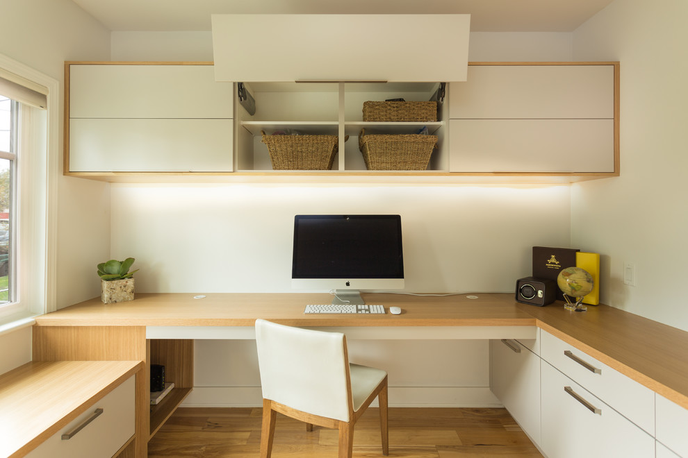 Ejemplo de despacho actual de tamaño medio con paredes blancas, suelo de madera clara, escritorio empotrado y suelo marrón