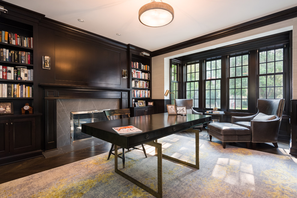 Exempel på ett klassiskt hemmabibliotek, med svarta väggar, mörkt trägolv, en standard öppen spis, ett fristående skrivbord och brunt golv