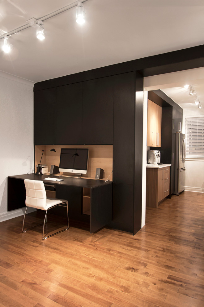 This is an example of a small contemporary home studio in Montreal with black walls, medium hardwood flooring, no fireplace and a built-in desk.
