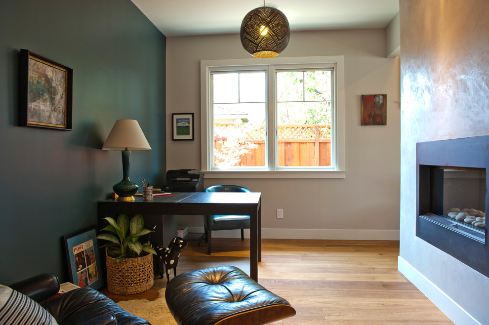 Foto de despacho actual pequeño con paredes multicolor, suelo de madera clara, chimenea lineal, marco de chimenea de metal y escritorio independiente