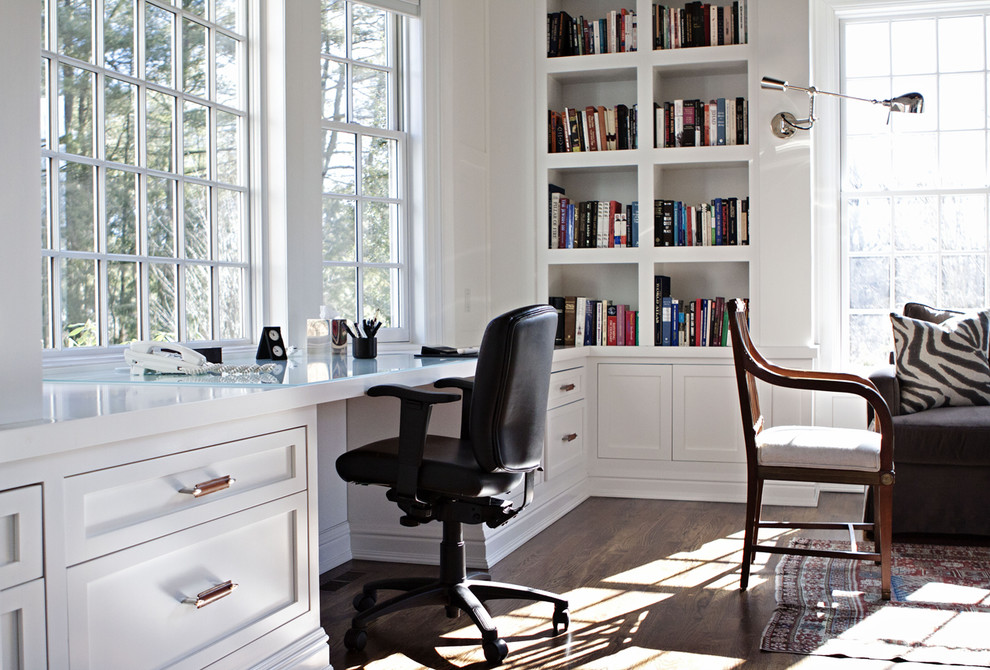 Modelo de despacho campestre extra grande sin chimenea con biblioteca, paredes blancas, suelo de madera oscura, escritorio empotrado y suelo marrón