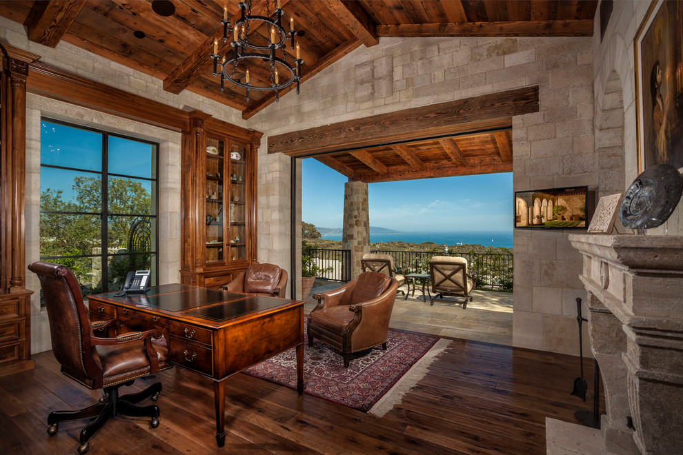 Inspiration for a mediterranean study in Orange County with beige walls, dark hardwood flooring, a standard fireplace, a freestanding desk and brown floors.