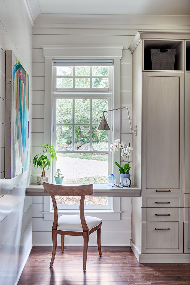 This is an example of a classic study in Charlotte with white walls, medium hardwood flooring, a built-in desk and brown floors.