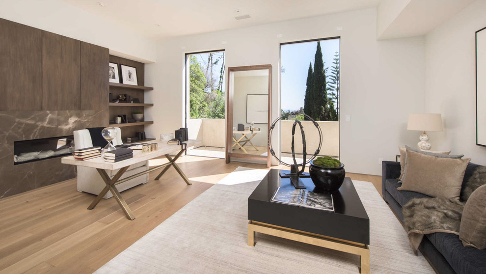 This is an example of a contemporary study in Los Angeles with white walls, light hardwood flooring, a ribbon fireplace, a stone fireplace surround, a freestanding desk and beige floors.
