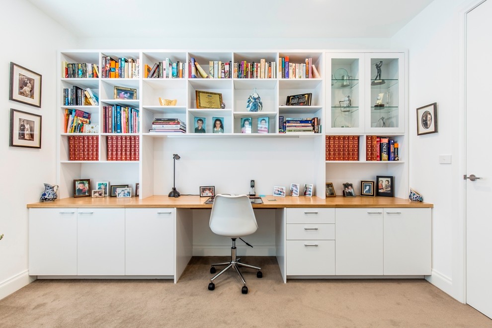 Bild på ett mellanstort funkis hemmabibliotek, med ett inbyggt skrivbord, vita väggar och heltäckningsmatta