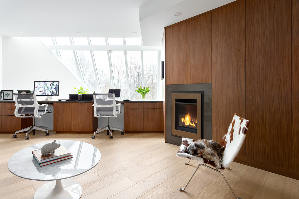 Photo of a contemporary home office in Vancouver with white walls, light hardwood flooring, a standard fireplace, a metal fireplace surround, a built-in desk and beige floors.