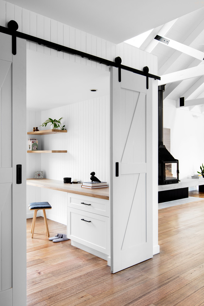 Cette image montre un petit bureau design avec un mur blanc, un sol en bois brun, un poêle à bois, un manteau de cheminée en béton, un bureau intégré, un plafond voûté et du lambris.