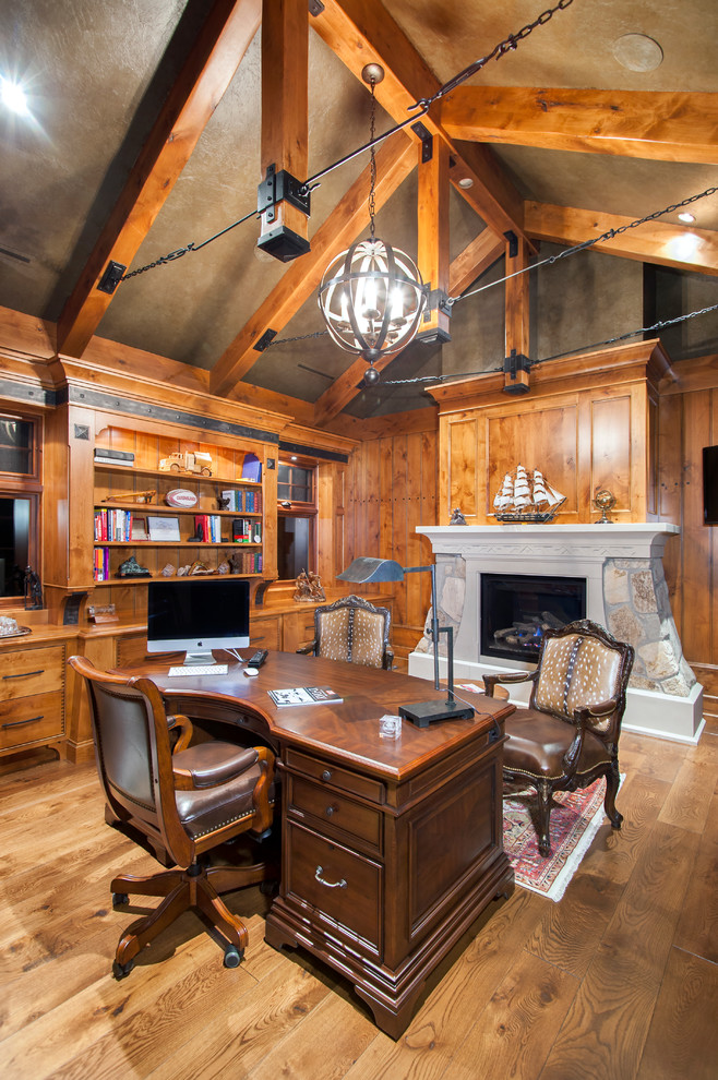 Foto de despacho rural grande con suelo de madera en tonos medios, todas las chimeneas, marco de chimenea de piedra y escritorio independiente