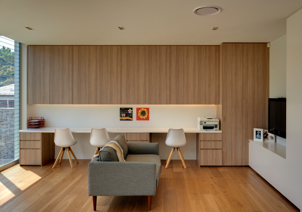 Modelo de despacho contemporáneo grande sin chimenea con paredes blancas, escritorio empotrado y suelo de madera en tonos medios