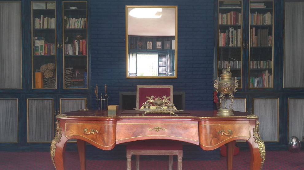 Inredning av ett klassiskt mellanstort arbetsrum, med ett bibliotek, blå väggar, heltäckningsmatta, en standard öppen spis, en spiselkrans i sten, ett fristående skrivbord och rött golv