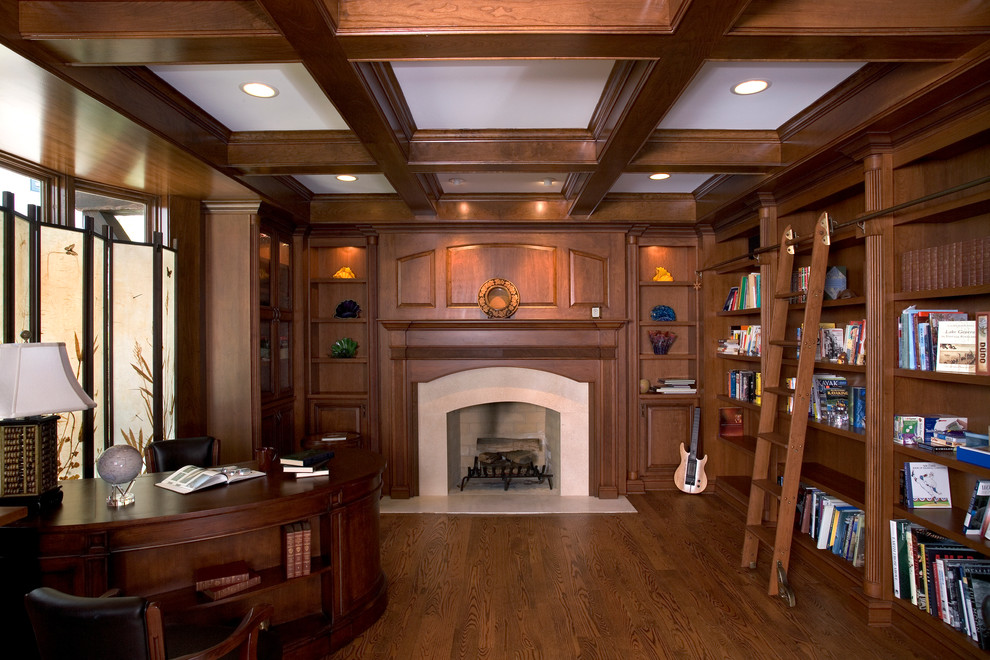 Inredning av ett klassiskt stort hemmabibliotek, med bruna väggar, mellanmörkt trägolv, en standard öppen spis, en spiselkrans i sten och ett inbyggt skrivbord