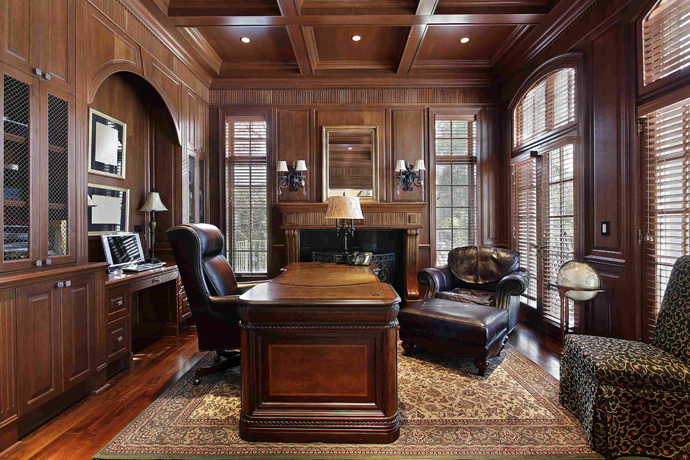 Klassisk inredning av ett mellanstort arbetsrum, med mörkt trägolv, en standard öppen spis, en spiselkrans i trä, ett fristående skrivbord, brunt golv och bruna väggar